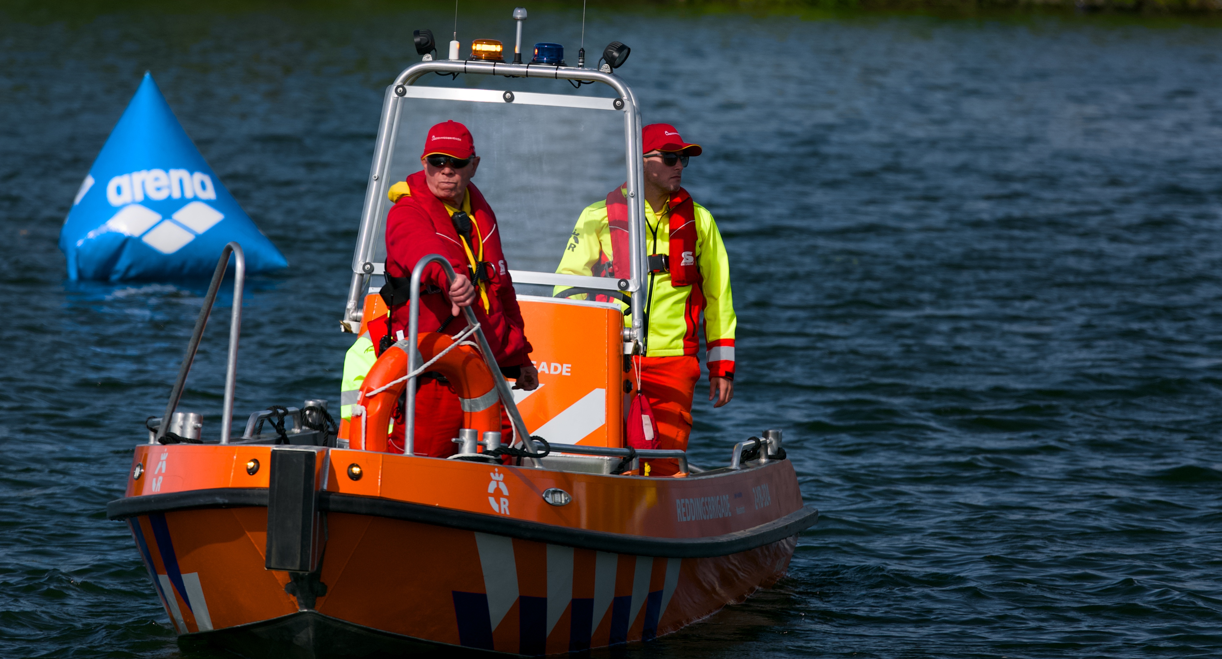 Een reddingsboot met twee lifeguards welke het water in de gaten houden. In de achtergrond drijft een driehoekige boei met het arena logo erop.